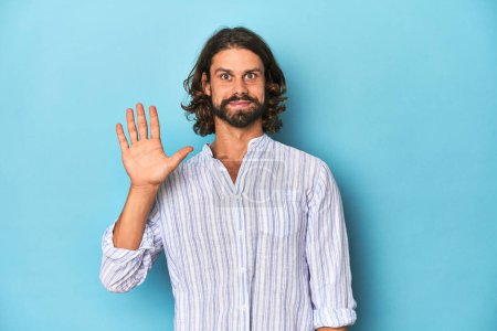 Photo for Man with beard in blue striped shirt, blue studio smiling cheerful showing number five with fingers. - Royalty Free Image