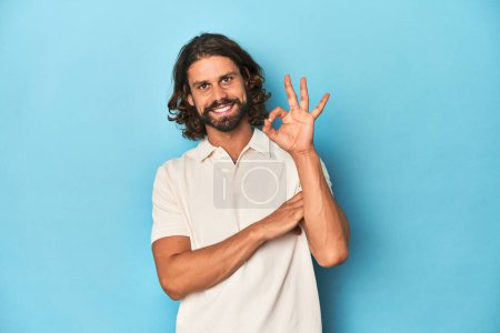 Téléchargez les photos : Homme aux cheveux longs dans un polo blanc, studio bleu claque des yeux et tient un geste correct avec la main. - en image libre de droit