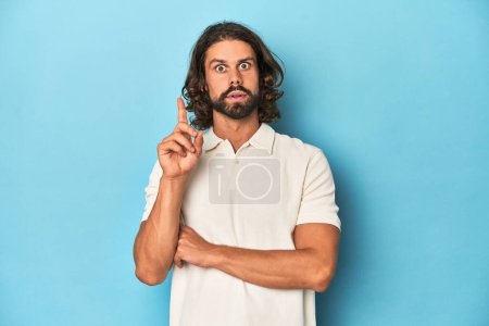 Téléchargez les photos : Homme aux cheveux longs dans un polo blanc, studio bleu ayant une bonne idée, concept de créativité. - en image libre de droit