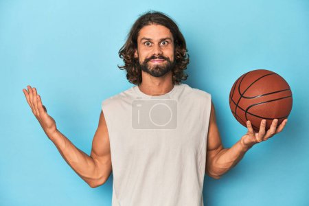 Téléchargez les photos : Homme barbu avec basket en studio bleu recevant une agréable surprise, excité et levant la main. - en image libre de droit