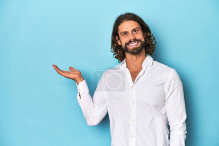 Photo for Bearded man in a white shirt, blue backdrop showing a copy space on a palm and holding another hand on waist. - Royalty Free Image