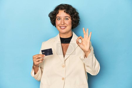 Photo for Young woman with credit card, elegant white blazer cheerful and confident showing ok gesture. - Royalty Free Image
