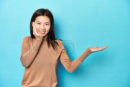 Téléchargez les photos : Jeune femme asiatique au col roulé marron, tient un espace de copie sur une paume, garde la main sur la joue. Incroyable et ravi. - en image libre de droit