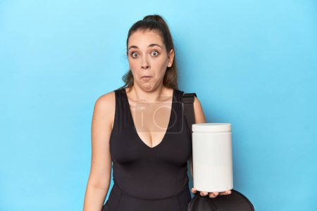 Foto de Mujer joven sosteniendo botella de proteína en ajuste deportivo encoge los hombros y los ojos abiertos confundido. - Imagen libre de derechos