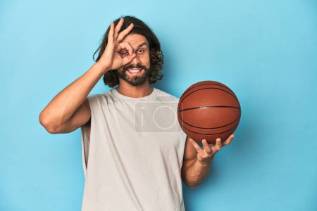 Photo for Bearded man with basketball in blue studio excited keeping ok gesture on eye. - Royalty Free Image