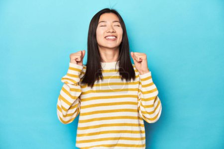 Téléchargez les photos : Femme asiatique en pull jaune rayé, célébrant une victoire, passion et enthousiasme, expression heureuse. - en image libre de droit