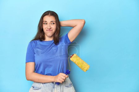 Téléchargez les photos : Jeune femme avec rouleau de peinture jaune sur un fond bleu touchant l'arrière de la tête, pensant et faisant un choix. - en image libre de droit