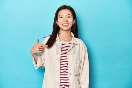 Téléchargez les photos : Femme asiatique en chemise à couches et t-shirt rayé, personne pointant à la main vers un espace de copie de chemise, fière et confiante - en image libre de droit