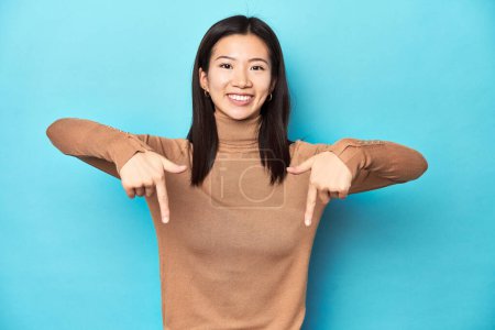 Téléchargez les photos : Jeune femme asiatique au col roulé marron, pointe vers le bas avec les doigts, sentiment positif. - en image libre de droit
