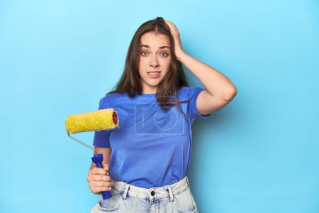 Photo for Young woman with yellow paint roller on a blue background being shocked, she has remembered important meeting. - Royalty Free Image