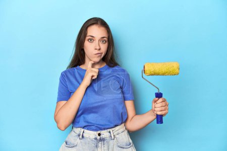 Foto de Joven mujer caucásica con rodillo de pintura amarillo en estudio azul. - Imagen libre de derechos