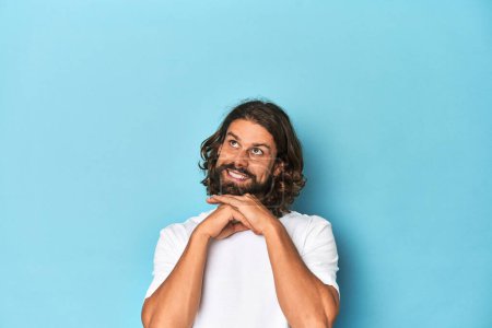Photo for Bearded man in a white shirt, blue backdrop keeps hands under chin, is looking happily aside. - Royalty Free Image