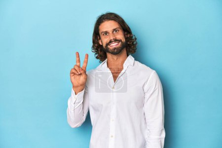 Photo for Bearded man in a white shirt, blue backdrop showing victory sign and smiling broadly. - Royalty Free Image