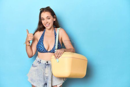 Foto de Mujer en bikini con un refrigerador de playa portátil, estudio azul sonriendo y levantando el pulgar hacia arriba - Imagen libre de derechos