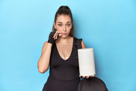 Photo for Young woman holding protein bottle in sporty setting with fingers on lips keeping a secret. - Royalty Free Image