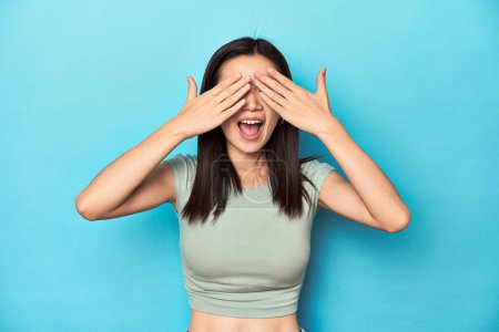 Photo for Asian woman in summer green top, studio backdrop, covers eyes with hands, smiles broadly waiting for a surprise. - Royalty Free Image