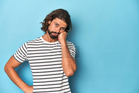 Téléchargez les photos : Homme barbu dans une chemise rayée, fond bleu qui se sent triste et coûteux, en regardant l'espace de copie. - en image libre de droit