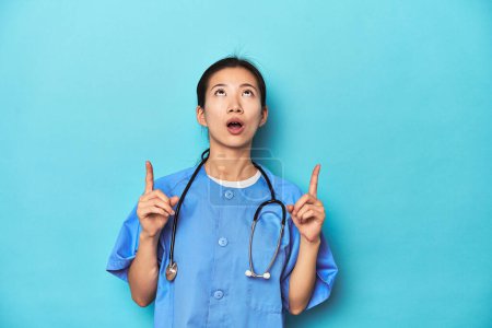 Photo for Asian nurse with stethoscope, medical studio shot, pointing upside with opened mouth. - Royalty Free Image