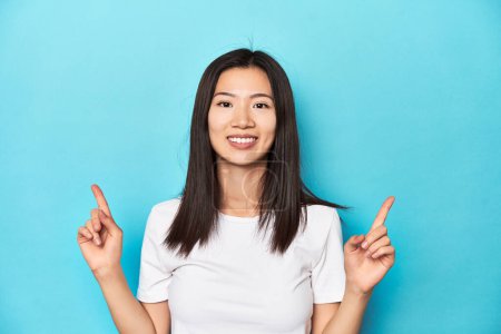 Foto de Mujer asiática joven en camiseta blanca, plano de estudio, indica con los dos dedos delanteros hacia arriba mostrando un espacio en blanco. - Imagen libre de derechos