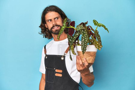 Photo for Long-haired gardener with beard holding a plant looks aside smiling, cheerful and pleasant. - Royalty Free Image