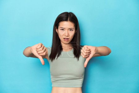 Téléchargez les photos : Asiatique femme en été vert top, studio toile de fond, montrant pouce vers le bas et exprimant dégoût. - en image libre de droit