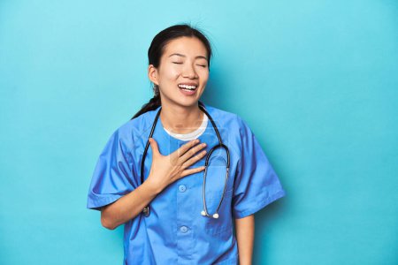 Téléchargez les photos : Infirmière asiatique avec stéthoscope, prise de vue en studio médical, rit fort en gardant la main sur la poitrine. - en image libre de droit