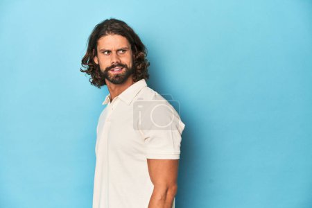 Téléchargez les photos : Homme aux cheveux longs dans un polo blanc, studio bleu regarde de côté souriant, gai et agréable. - en image libre de droit