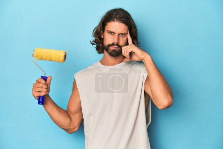 Photo for Bearded man painting with a yellow roller pointing temple with finger, thinking, focused on a task. - Royalty Free Image
