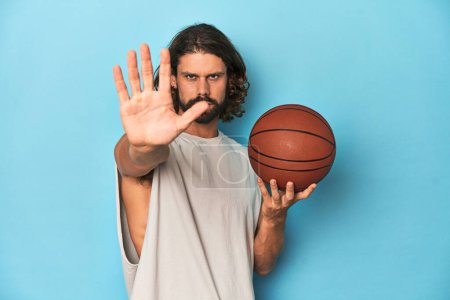 Téléchargez les photos : Homme barbu avec basket-ball en studio bleu debout avec la main tendue montrant le panneau d'arrêt, vous empêchant. - en image libre de droit