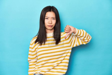 Téléchargez les photos : Femme asiatique en pull jaune rayé, montrant pouce baissé, concept de déception. - en image libre de droit
