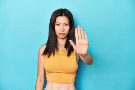 Téléchargez les photos : Femme asiatique en haut jaune d'été, installation du studio, debout avec la main tendue montrant le panneau stop, vous empêchant. - en image libre de droit