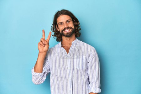 Foto de Hombre con barba en camisa de rayas azules, estudio azul mostrando el número dos con los dedos. - Imagen libre de derechos