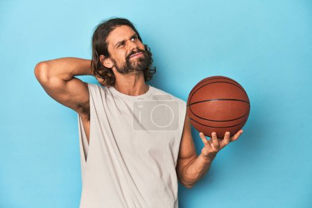 Foto de Hombre barbudo con baloncesto en estudio azul tocando la parte posterior de la cabeza, pensando y haciendo una elección. - Imagen libre de derechos