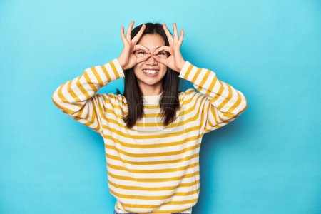 Photo for Asian woman in striped yellow sweater, showing okay sign over eyes - Royalty Free Image
