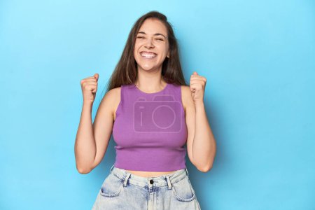 Téléchargez les photos : Jeune femme à la mode dans un haut violet sur fond bleu célébrant une victoire, passion et enthousiasme, expression heureuse. - en image libre de droit