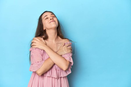 Photo for Young woman wearing a pink dress on a blue studio backdrop hugs, smiling carefree and happy. - Royalty Free Image