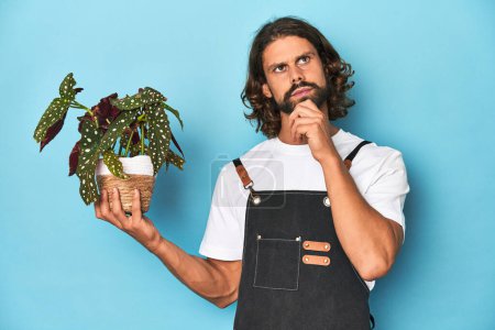 Foto de Jardinero de pelo largo con barba sosteniendo una planta mirando hacia los lados con expresión dudosa y escéptica. - Imagen libre de derechos