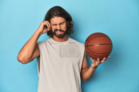 Foto de Hombre barbudo con baloncesto en estudio azul cubriendo orejas con las manos. - Imagen libre de derechos