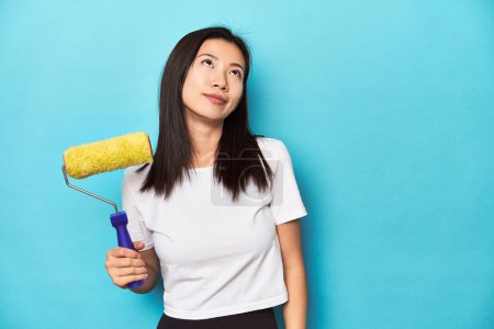 Téléchargez les photos : Jeune femme asiatique avec rouleau de peinture, concept de bricolage, rêvant d'atteindre des objectifs et des buts - en image libre de droit