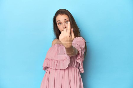 Téléchargez les photos : Jeune femme vêtue d'une robe rose sur fond de studio bleu pointant du doigt vers vous comme si elle vous invitait à vous approcher. - en image libre de droit