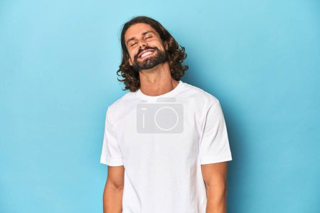 Foto de Hombre barbudo con camisa blanca, fondo azul se ríe y cierra los ojos, se siente relajado y feliz. - Imagen libre de derechos