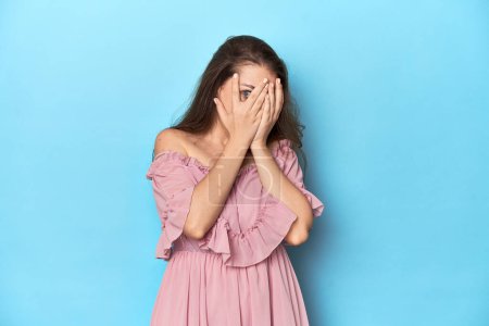 Téléchargez les photos : Jeune femme portant une robe rose sur fond bleu studio cligner des yeux à travers les doigts effrayé et nerveux. - en image libre de droit