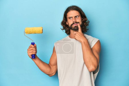Photo for Bearded man painting with a yellow roller relaxed thinking about something looking at a copy space. - Royalty Free Image