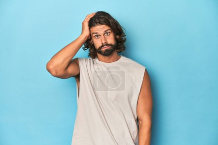 Photo for Bearded man in a tank top, blue backdrop being shocked, she has remembered important meeting. - Royalty Free Image