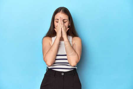 Photo for Woman in a striped top posing on a blue studio backdrop holding hands in pray near mouth, feels confident. - Royalty Free Image
