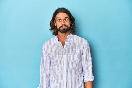 Photo for Man with beard in blue striped shirt, blue studio shrugs shoulders and open eyes confused. - Royalty Free Image