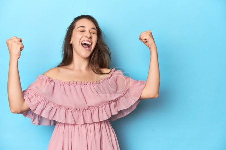 Photo for Young woman wearing a pink dress on a blue studio backdrop raising fist after a victory, winner concept. - Royalty Free Image
