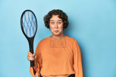 Photo for Young woman with electric mosquito racket Young woman with electric mosquito racketshrugs shoulders and open eyes confused. - Royalty Free Image
