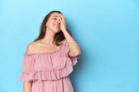 Téléchargez les photos : Jeune femme portant une robe rose sur fond de studio bleu riant heureux, insouciant, émotion naturelle. - en image libre de droit