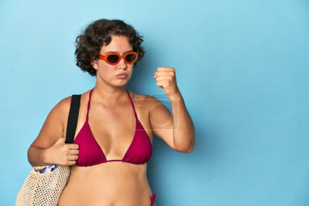 Photo for Young woman in bikini with beach bag showing fist to camera, aggressive facial expression. - Royalty Free Image